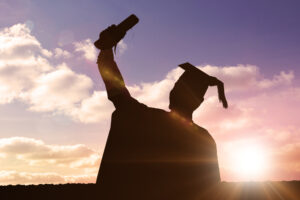 The sunset casts a silhouette of a college student wearing a cap and gown, raising their diploma into the air.