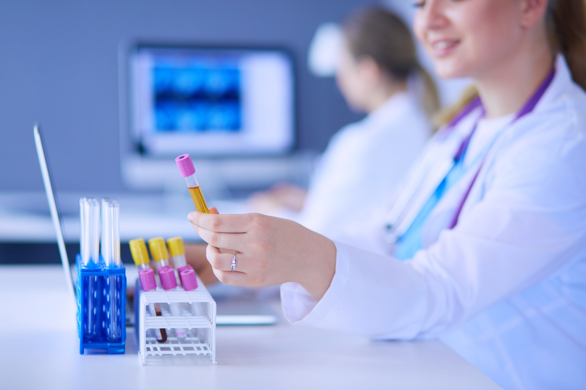 A technician in a white lab coat holds a test tube above a small rack containing several additional test tubes filled with various fluids.