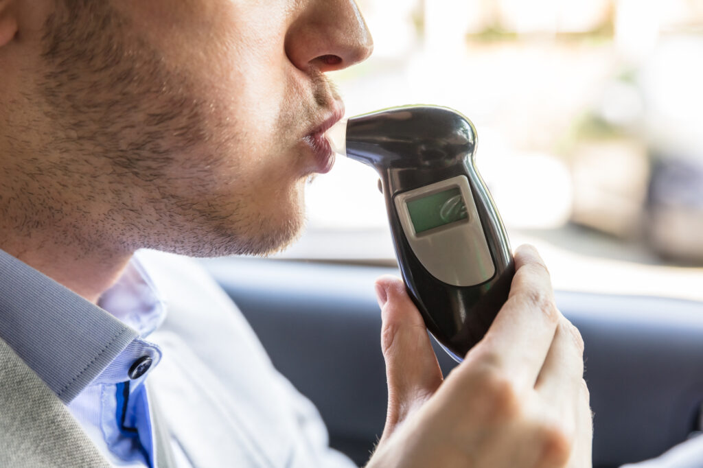 Close-up of a person seated in a car, blowing into a digital alcohol breathalyzer device.