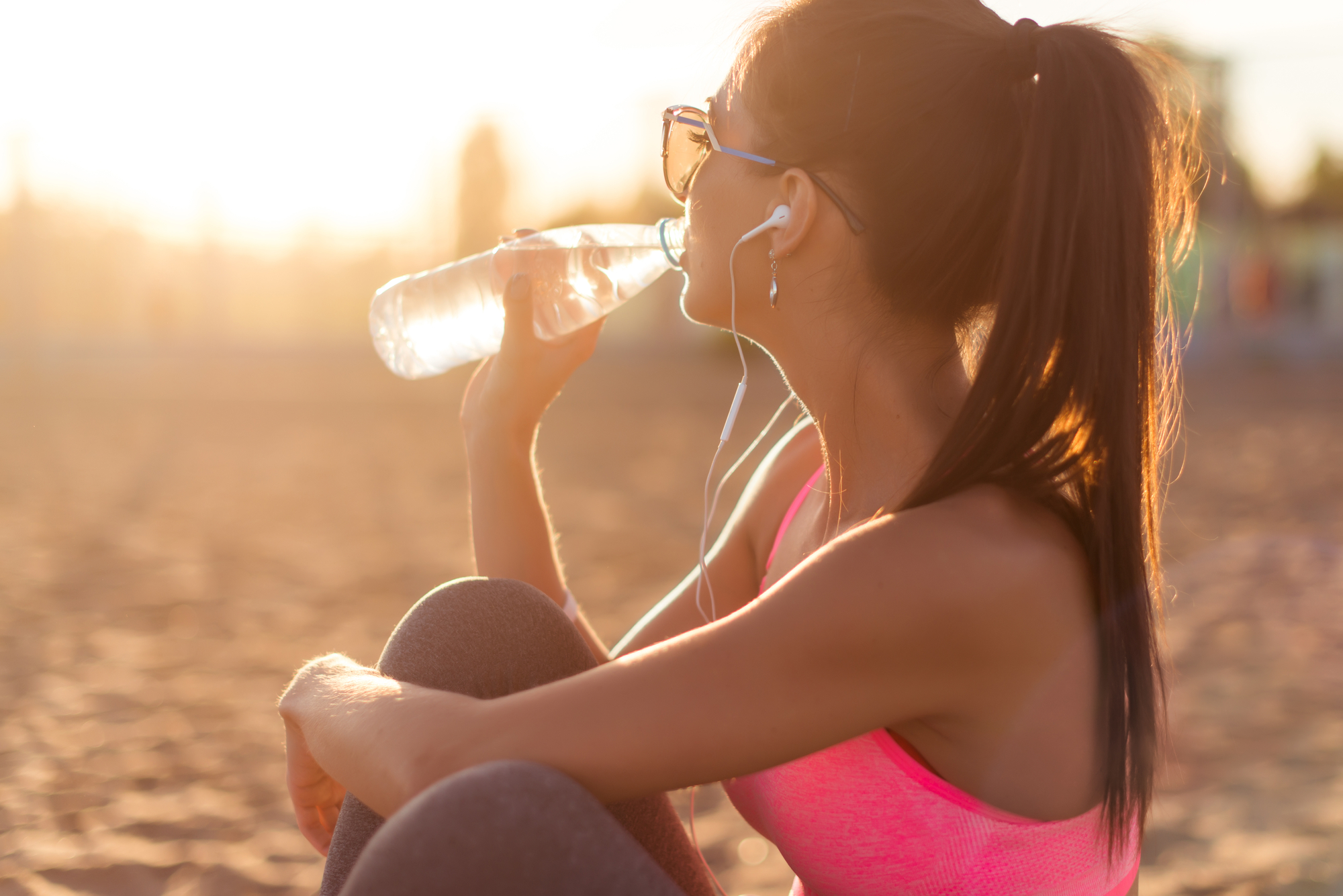 An individual in athletic apparel sitting in the sand at the beach at sunset, listening to music through earbuds and drinking bottled water.