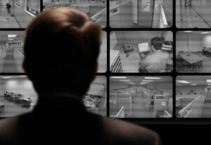 Over-the-shoulder view from behind a person in shadows observing an office via closed-circuit televisions and cameras.