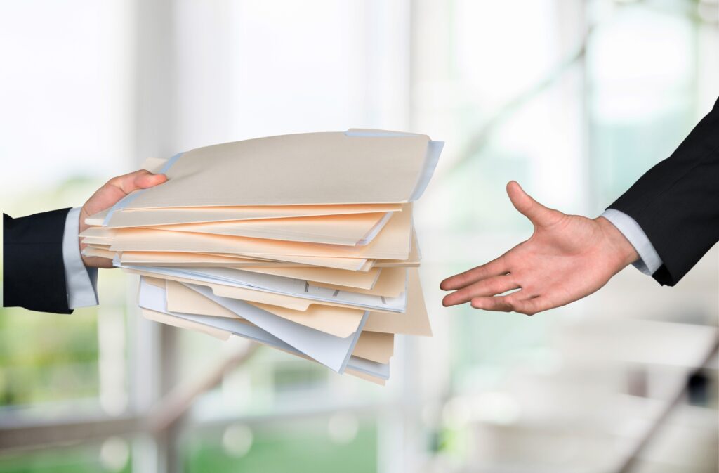 Close-up of a person’s hand holding a large stack of folders, passing them to another person wearing a suit.