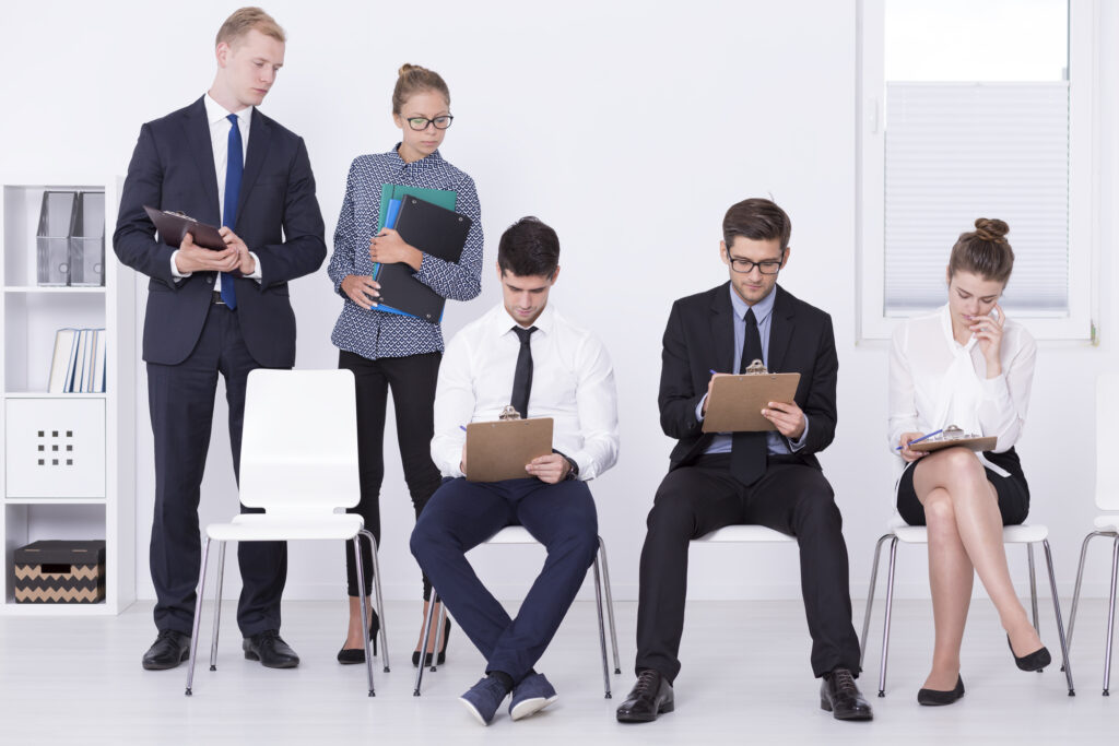 A row of individuals wearing business apparel seated and filling out forms on clipboards, while two other people watch over them.