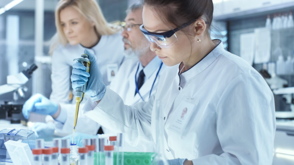 Three scientists in white lab coats and goggles working with vials and a pipette.