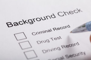 Close-up of a ballpoint pen hovering over a document labeled “Background Check” with empty checkboxes available for “Criminal Record,” “Drug Test,” Driving Record,” and “Social Security” visible.